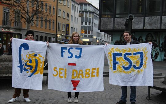 Drei Menschen halten bemalte Tücher in die Luft mit der Aufschrift 